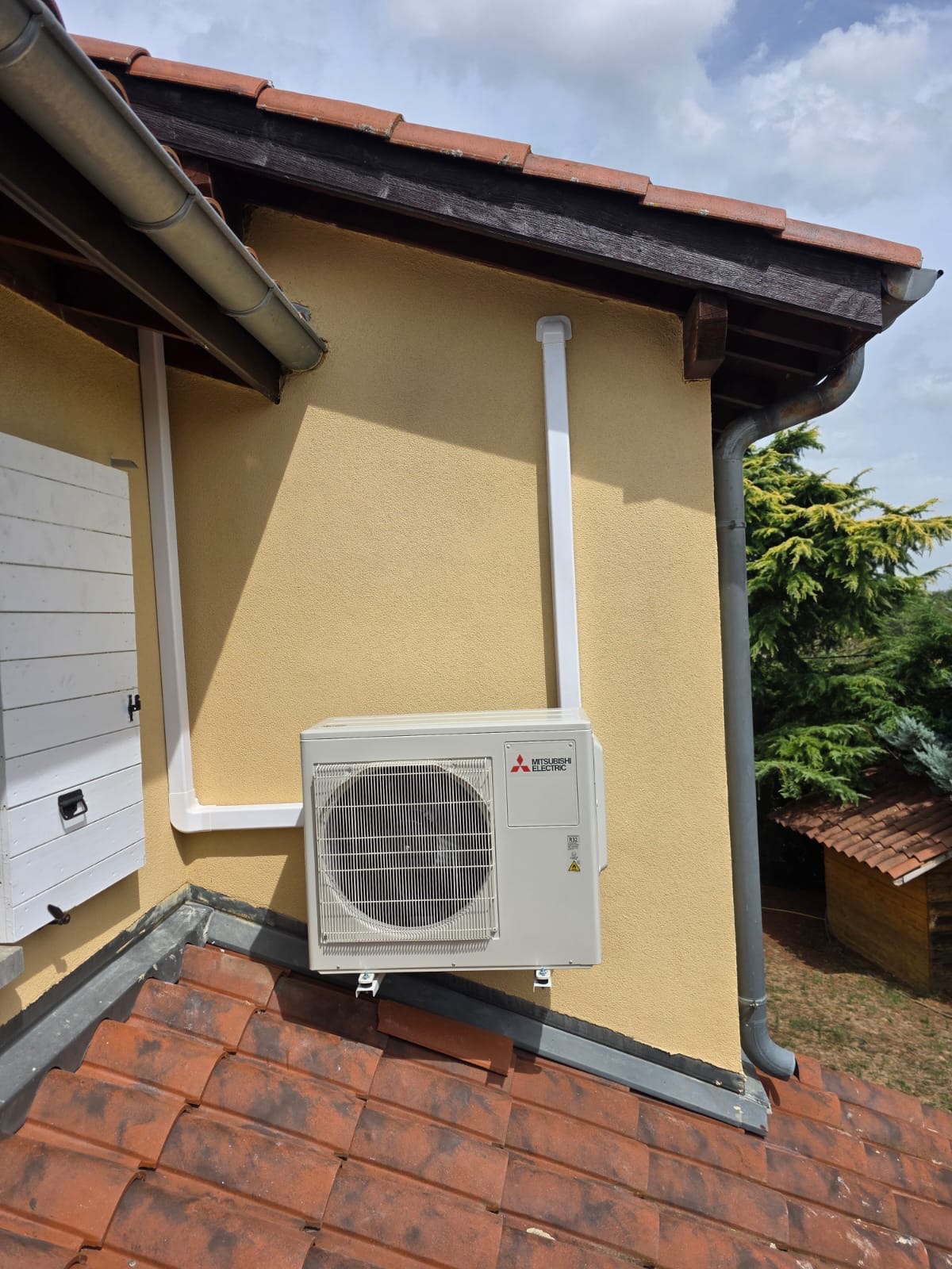 Unité extérieure de climatisation posée sur supports en toiture, le long d’une façade crépie