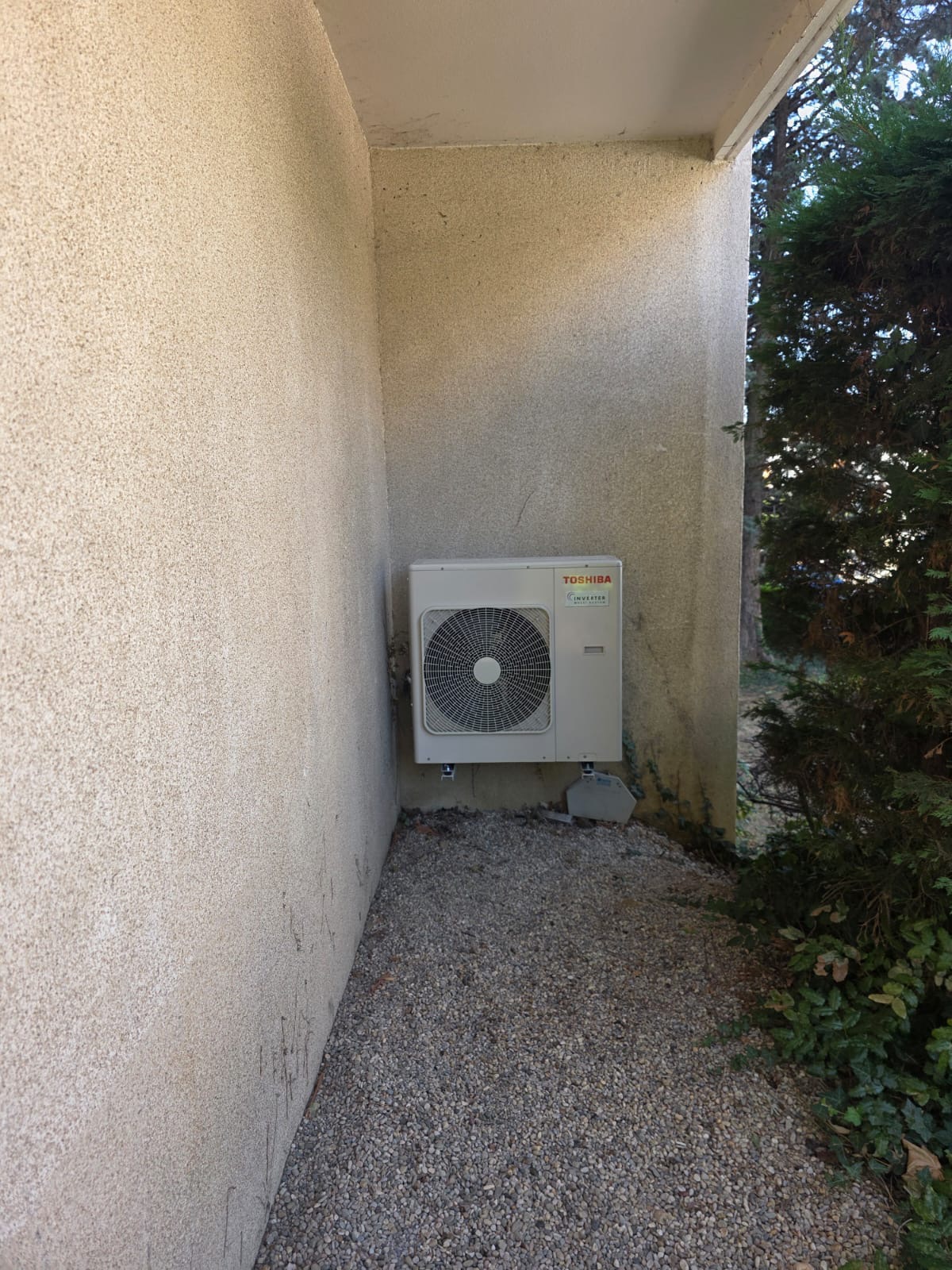 Unité extérieure de climatisation fixée sur consoles dans un passage étroit, sol en gravier